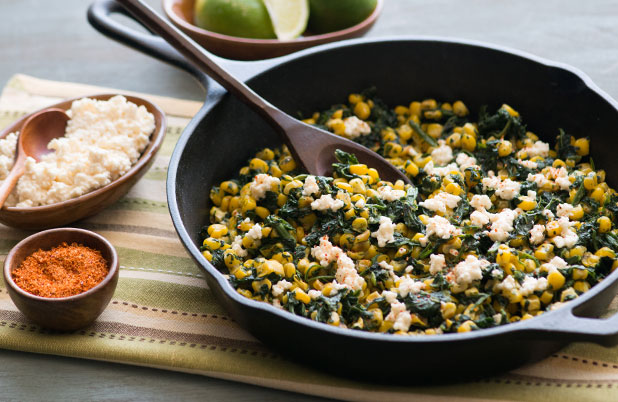 A skillet with corn, spinach and cotija cheese on a napkin beside bowls of cheese and cayenne.